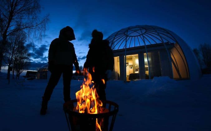 Due persone di un viaggio di gruppo WeRoad si stagliano contro un falò nella neve al tramonto, con un igloo di vetro sullo sfondo.