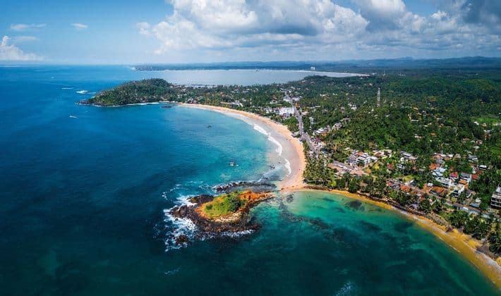 An aerial view of a tropical coastline with a crescent beach, a rocky islet, and turquoise water under a partly cloudy sky.