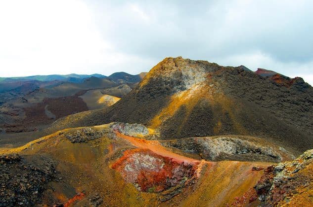 A colorful volcanic landscape with craters and dark rocky hills under a cloudy sky.