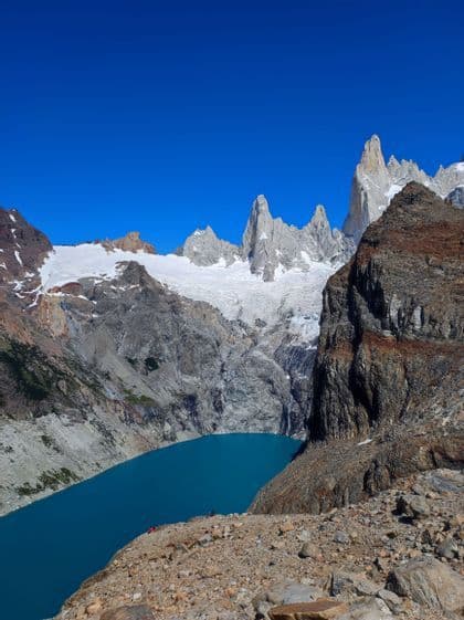 Montagne frastagliate e innevate e un grande ghiacciaio dominano un lago alpino turchese sotto un cielo limpido e azzurro.