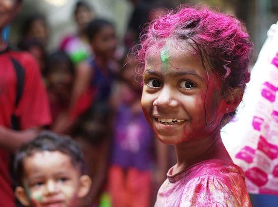 Una niña sonriente con el pelo rosa brillante y polvo de colores en la cara mira a la cámara durante una celebración.