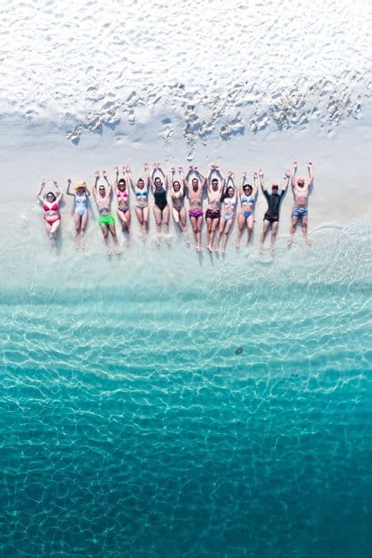 An aerial view of a WeRoad group trip lying in a row on a white sand beach with their arms raised, feet in the clear turquoise water.