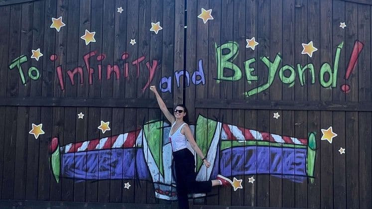 Eine lächelnde Frau posiert verspielt vor einer Holzwand mit dem Graffiti 'Ins Unendliche und darüber hinaus!'