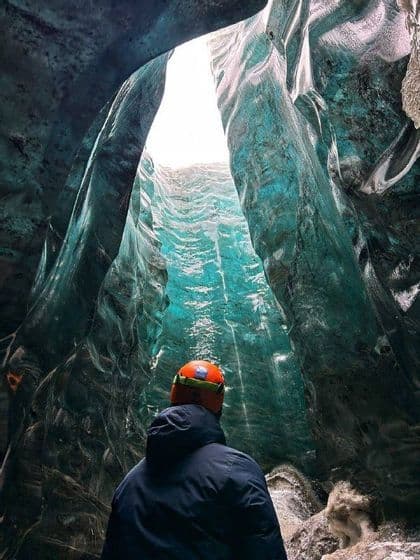 Una persona con un casco arancione si trova all'interno di una grotta di ghiaccio blu, guardando verso un'apertura nel cielo.