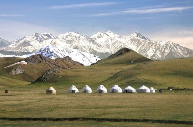 A row of yurts sits on a wide green plain, with rolling hills and a vast, snow-capped mountain range behind them.