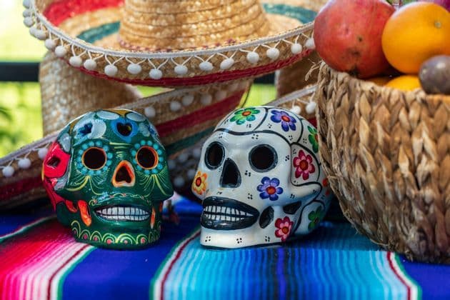 Dos calaveras de azúcar coloridas reposan sobre un serape a rayas junto a una cesta de frutas y sombreros.