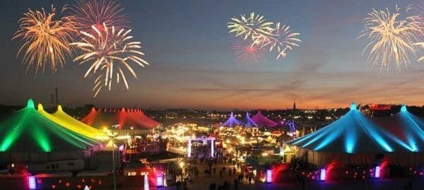 Fuochi d'artificio esplodono su un festival affollato con grandi tende colorate e illuminate al crepuscolo.