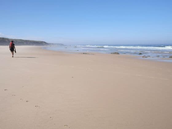 Una persona con un grande zaino cammina a piedi nudi su un'ampia spiaggia sabbiosa, lasciandosi dietro delle impronte.