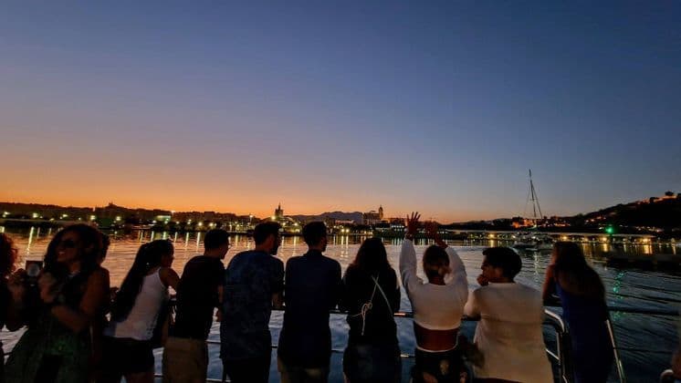 Un gruppo WeRoad si affaccia dal parapetto di una barca, ammirando lo skyline illuminato di una città sull'acqua al crepuscolo.