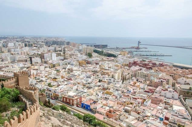 Una vista aerea di una città costiera densamente popolata, con un'antica cinta muraria fortificata in primo piano e un grande porto sullo sfondo.