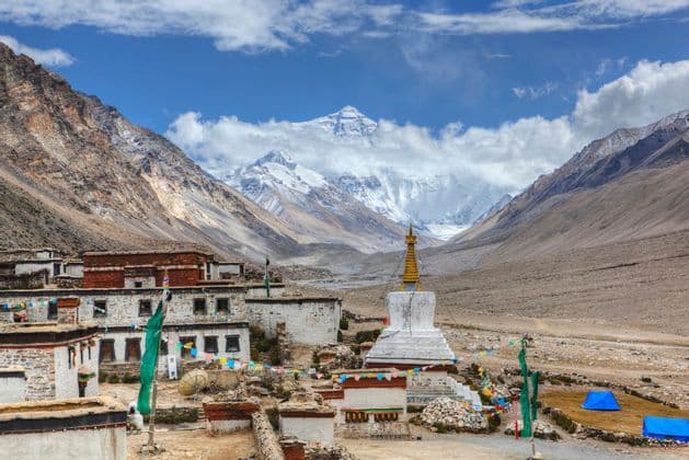 Un pueblo con una estupa blanca y banderas de oración se asienta en un valle de montaña, con un gran pico cubierto de nieve elevándose al fondo.