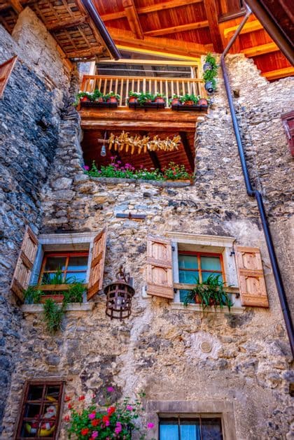Una vista dal basso di un edificio rustico in pietra con persiane in legno, balconi fioriti e pannocchie di mais essiccate appese