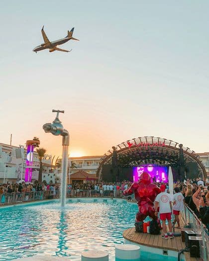 Un aereo sorvola un affollato festival all'aperto con una piscina, un grande palco e una fontana a forma di rubinetto al tramonto.