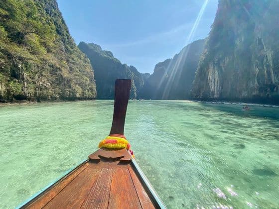 Una vista dalla prua di una barca tradizionale a coda lunga in legno che solca acque cristalline color turchese in una laguna circondata da alte scogliere verdi.