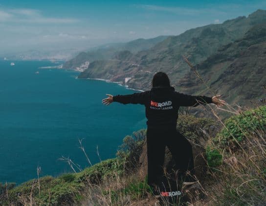 Un líder de grupo de WeRoad de pie con los brazos extendidos en un acantilado cubierto de hierba, contemplando un vasto mar azul y una costa montañosa.