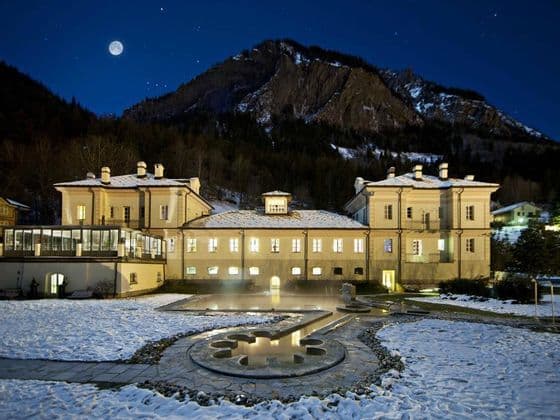 Un edificio illuminato con una piscina termale esterna in un paesaggio innevato, con una grande montagna e una luna piena nel cielo notturno.