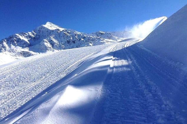 Una pista da sci battuta con tracce che conducono verso montagne innevate sotto un cielo azzurro brillante.