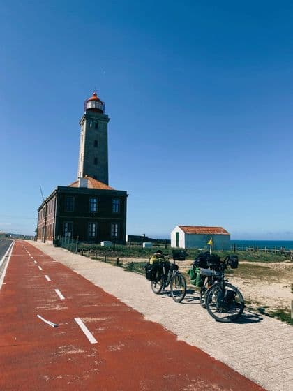 Due biciclette da turismo cariche di attrezzatura sono parcheggiate su un sentiero costiero rosso, con un alto faro in pietra sullo sfondo.