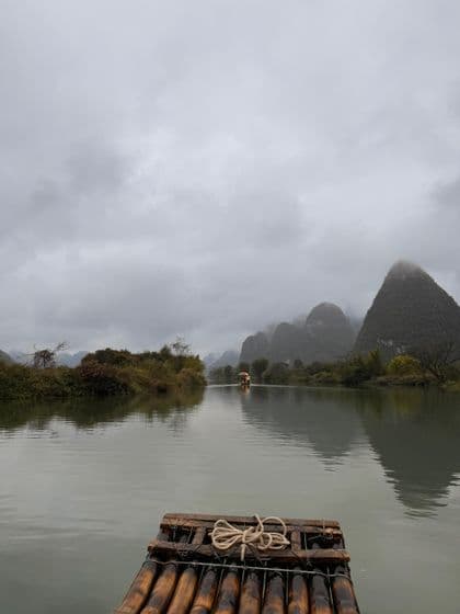 Vista dalla parte anteriore di una zattera di bambù su un fiume calmo, con montagne carsiche avvolte nella nebbia sotto un cielo pesante e coperto.