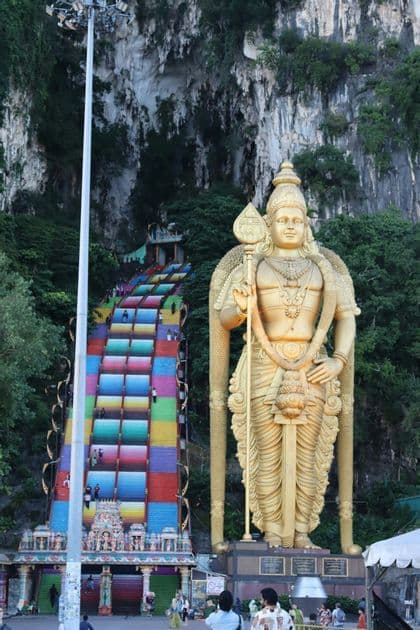 Eine riesige goldene Statue einer Gottheit steht neben einer langen, farbenfrohen Treppe, die eine felsige Klippe hinaufführt, mit Menschen auf den Stufen.