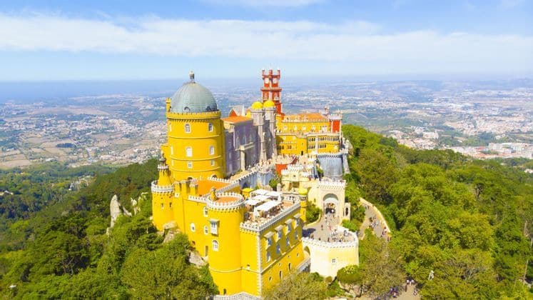 An aerial view of a colorful yellow and red castle perched on a lush, forested hilltop overlooking a sprawling city and the ocean.
