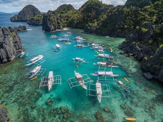 An aerial view of numerous outrigger boats and kayaks floating in a clear turquoise lagoon surrounded by steep, jungle-covered cliffs.