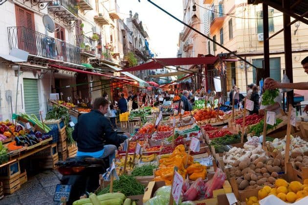 Un vivace mercato di strada all'aperto con bancarelle piene di frutta e verdura colorata mentre le persone curiosano e fanno acquisti.