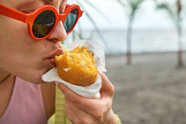 Primo piano di una persona che indossa occhiali da sole rossi mentre mangia un arancino fritto ripieno di formaggio su una spiaggia con palme sullo sfondo.