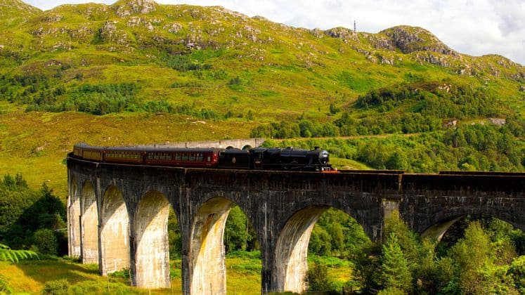 Ein schwarzer Dampfzug mit roten Waggons fährt über ein steinernes Viadukt, mit einem üppig grünen Berg im Hintergrund.