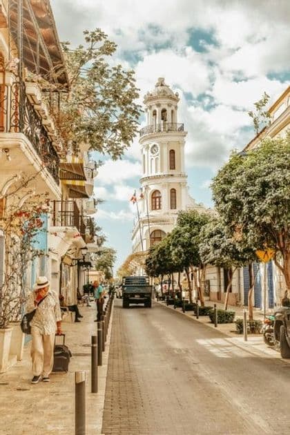 A narrow, tree-lined city street with historic buildings leads towards a white clock tower under a partly cloudy sky.