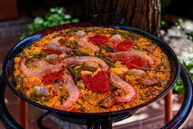 Eine Paella-Pfanne mit gelbem Reis, Garnelen, roten Paprika und grünen Bohnen steht im Freien auf einem Ständer.