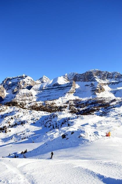 A person skis down a wide, sunny slope against a backdrop of snow-covered mountains under a clear blue sky.