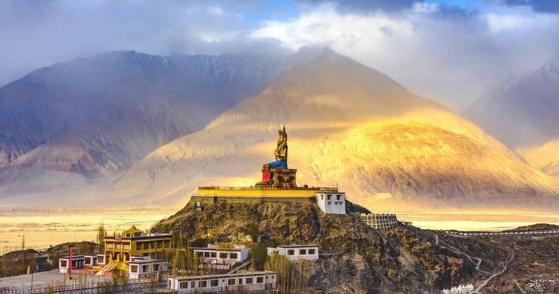 Une grande statue de Bouddha assise, dans un monastère perché au sommet d'une colline, surplombe une vallée avec de vastes montagnes en arrière-plan.