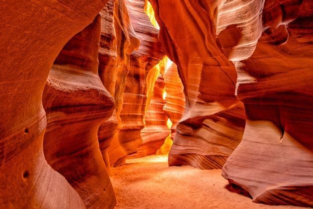 La luce del sole illumina il fondo sabbioso di uno stretto canyon a fessura con pareti rocciose ondulate, stratificate, rosse e arancioni.