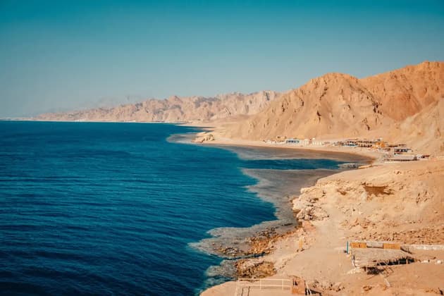 A panoramic view of a deep blue sea meeting a rugged, arid coastline with mountains under a clear sky.