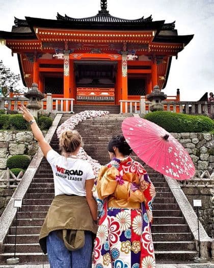 Un accompagnateur WeRoad se tient aux côtés d'une femme en kimono coloré, au pied de marches en pierre menant à un temple japonais orange.