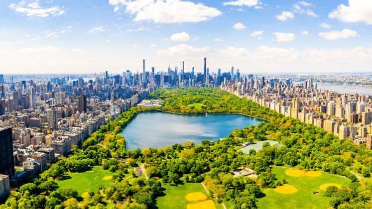 Eine Luftaufnahme eines großen Stadtparks mit einem zentralen See, umgeben von Wolkenkratzern und Gebäuden an einem sonnigen Tag.
