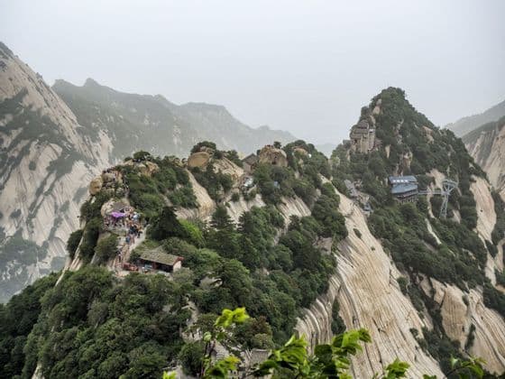 Una vista de una escarpada cordillera con picos rocosos, donde multitudes de personas caminan por un sendero entre templos y árboles verdes.