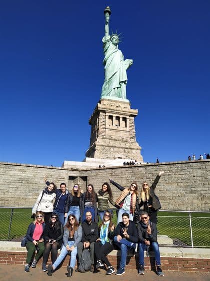 Eine WeRoad Gruppenreise posiert an einem sonnigen Tag für ein Foto vor einer großen grünen Statue einer Frau, die eine Fackel hält.