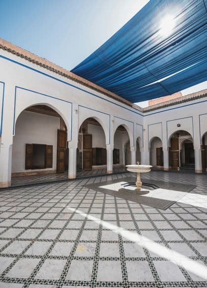 Un cortile soleggiato con una fontana centrale, pavimenti piastrellati geometrici e archi bianchi, sotto una grande tenda parasole blu.