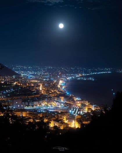Una luna piena pende nel cielo scuro sopra una vasta città costiera illuminata da luci gialle e blu di notte, vista da un punto panoramico elevato.