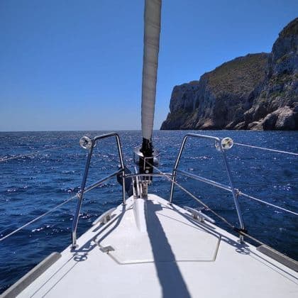 Vista desde la proa de un velero navegando por el mar azul hacia un acantilado rocoso.