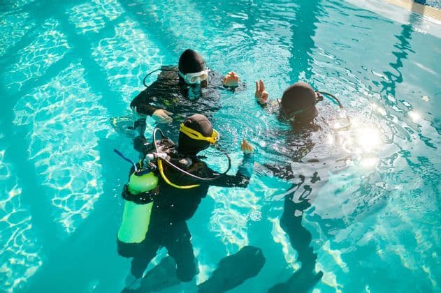 Trois participants d'un voyage en groupe WeRoad, équipés de matériel de plongée, s'exercent aux signaux manuels pendant un cours en piscine.