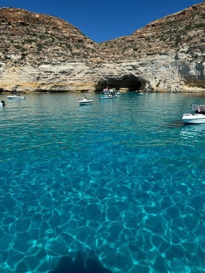 Barche su acque limpide e turchesi si avvicinano a una grotta marina alla base di una scogliera rocciosa stratificata sotto un cielo azzurro.