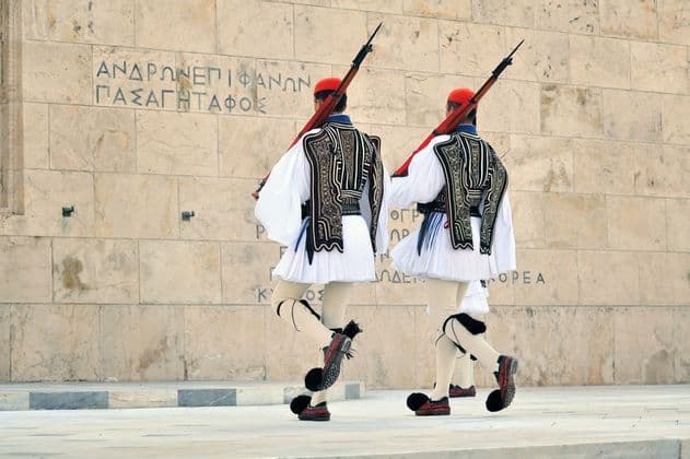 Dos guardias con uniformes griegos tradicionales, rifles y zapatos con pompones marchan al unísono frente a un muro de piedra con inscripciones.