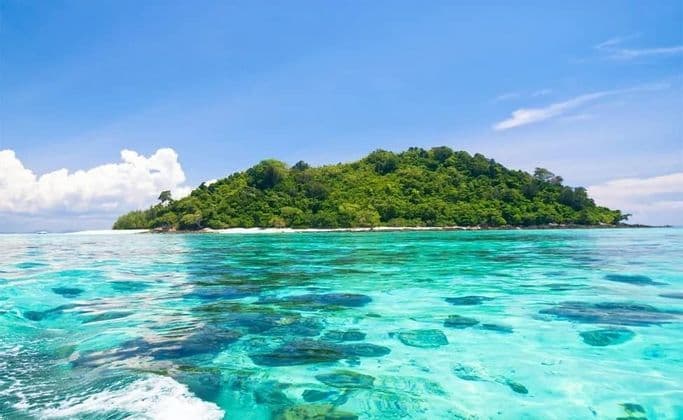 Una isla tropical cubierta de follaje verde, rodeada de agua clara turquesa bajo un cielo azul con nubes blancas.