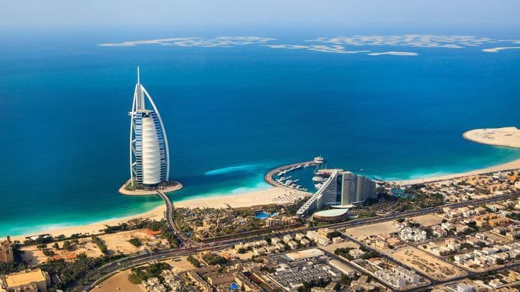 Veduta aerea di un hotel a forma di vela su una piccola isola artificiale, collegata da un ponte a una costa sabbiosa e a una città.