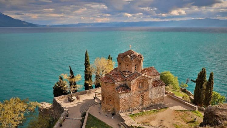 Una storica chiesa in pietra con il tetto di tegole rosse si erge su una scogliera che domina un vasto specchio d'acqua turchese sotto un cielo nuvoloso.