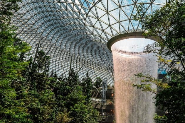 Ein Indoor-Wasserfall strömt aus einem Oberlicht in einer großen, geometrischen Glaskuppel, umgeben von üppigen grünen Bäumen und Laub.