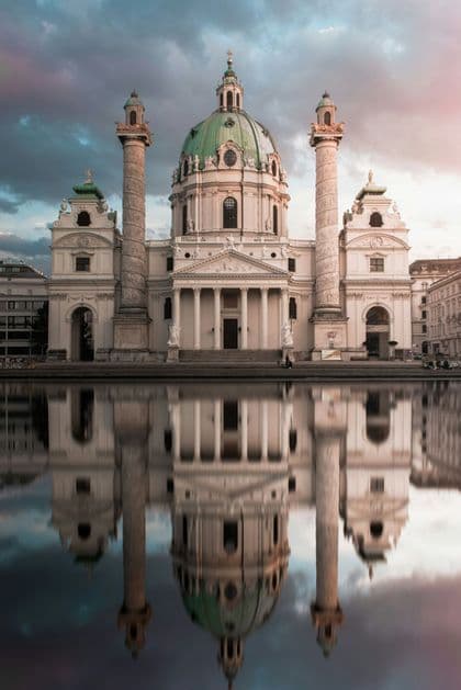 An ornate white church with a large green dome is perfectly reflected in the water before it under a cloudy sky.